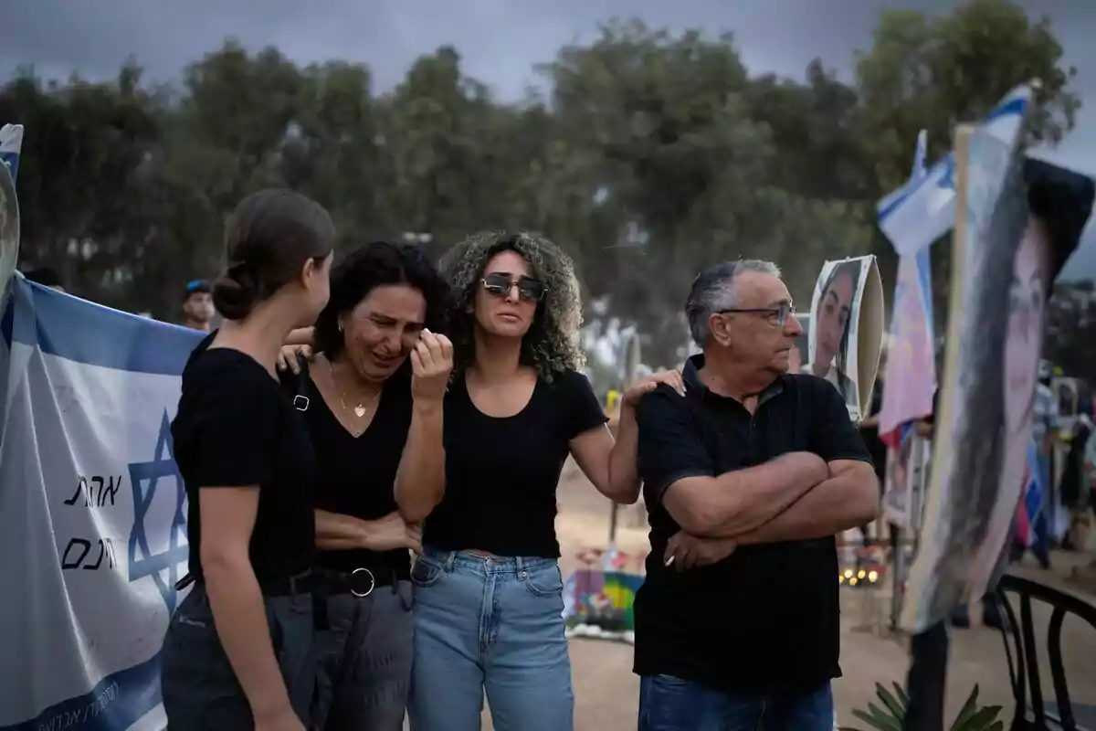 Un grup de persones vestides de negre es consolen mútuament en un ambient exterior amb banderes i fotografies al voltant. Un grup de persones vestides de negre es consolen mútuament en un ambient exterior amb banderes i fotografies al voltant.