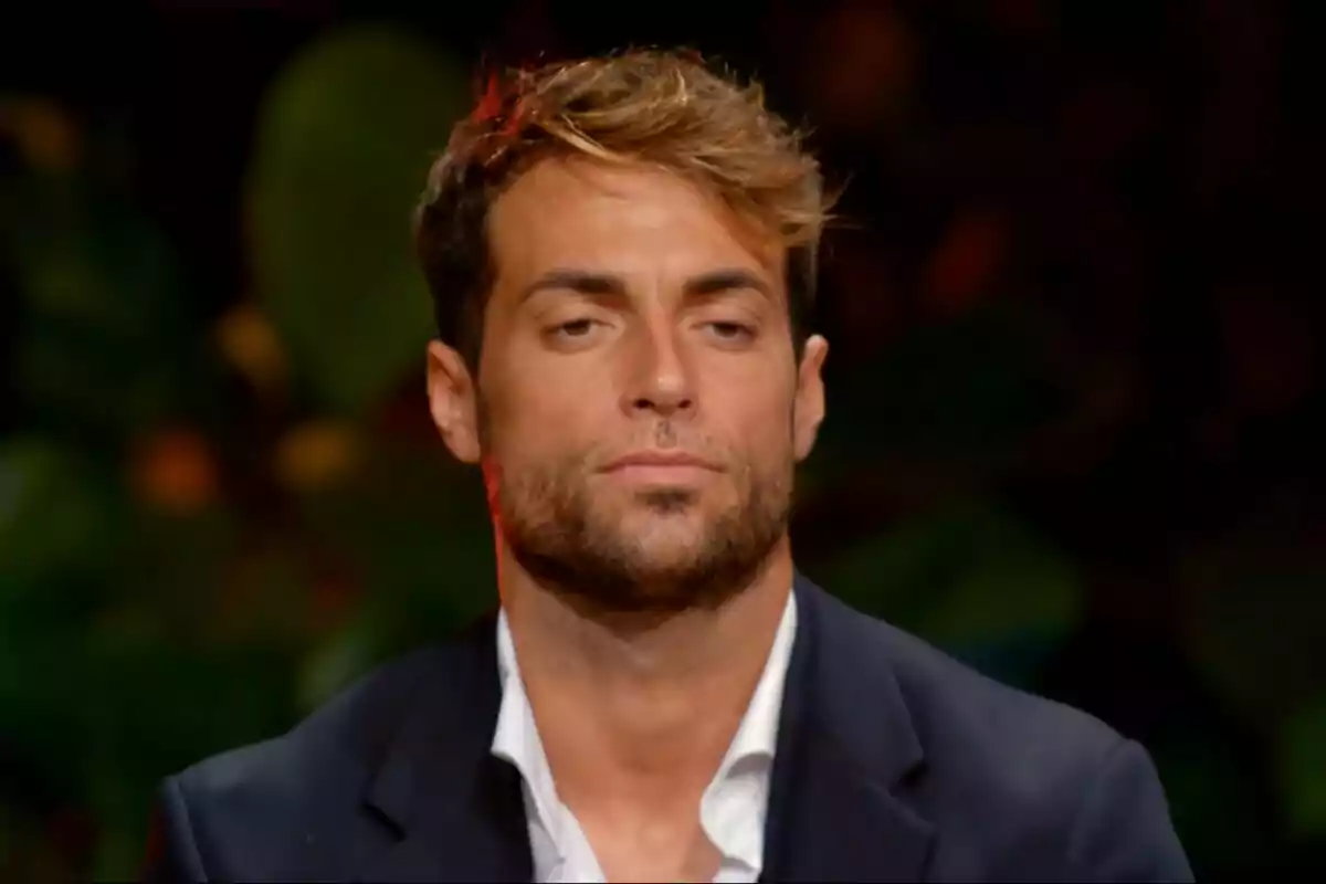 Montoya with a light brown beard and hair, wearing a dark suit and white jersey, with a serious expression and a blurred background on La Isla de las Tentaciones. Montoya with a light brown beard and hair, wearing a dark suit and white jersey, with a serious expression and a blurred background on La Isla de las Tentaciones.